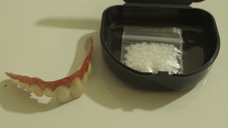 May include: A set of fake teeth in a plastic case with a small bag of adhesive. The teeth are made of a white plastic and have a pink gum line. The adhesive is in a clear plastic bag.