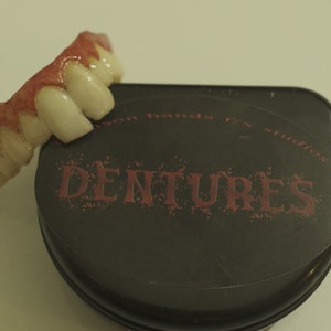 May include: A set of fake teeth with red gum detail, in a black case with the words "Dentures" printed in red.