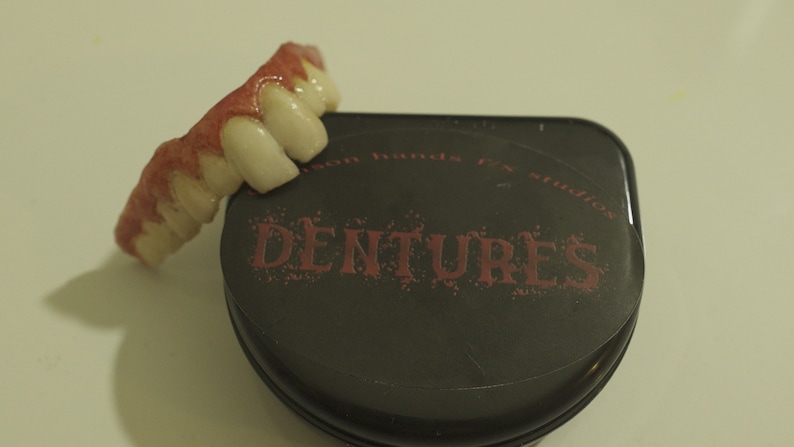 May include: A set of fake teeth with red gum detail, in a black case with the words "Dentures" printed in red.