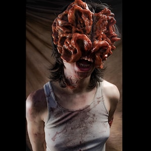 May include: A prosthetic mask featuring a complex, fleshy, fungal-like growth covering the upper face and eyes, with a visible mouth and teeth below. The figure wears a light-colored, ribbed tank top stained with dark, reddish-brown marks.