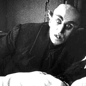 Pu&ograve; includere: Una fotografia in bianco e nero di un uomo con la pelle pallida e denti aguzzi, sdraiato a letto. Ha un'espressione preoccupata sul viso e gli occhi sono spalancati.