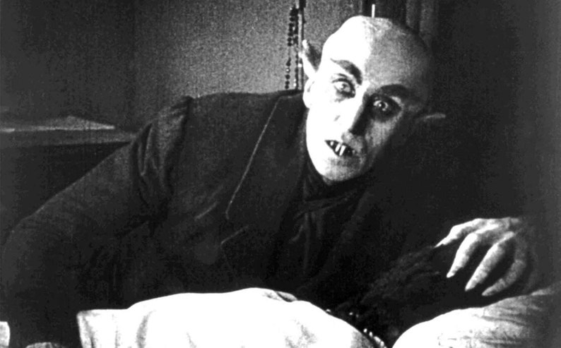 Pu&ograve; includere: Una fotografia in bianco e nero di un uomo con la pelle pallida e denti aguzzi, sdraiato a letto. Ha un'espressione preoccupata sul viso e gli occhi sono spalancati.