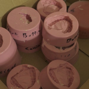 May include: A collection of pink silicone molds for making various shapes. The molds are stacked and arranged in a cardboard box. Some of the molds have handwritten labels.