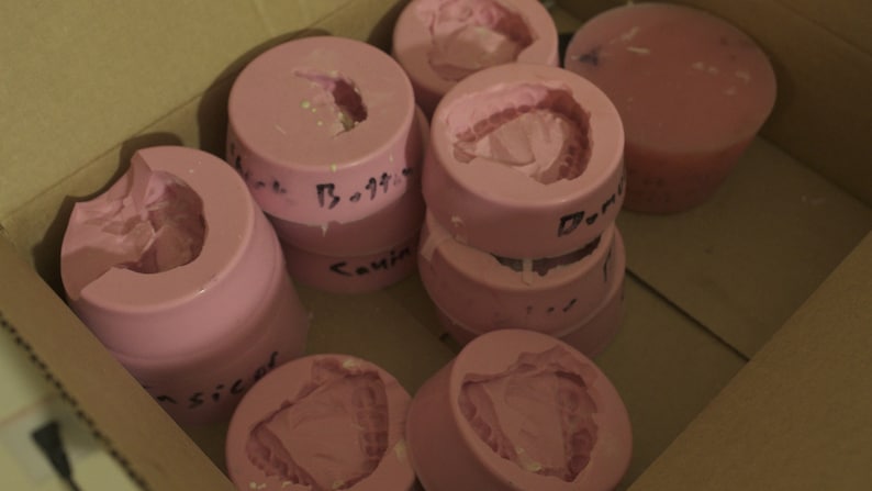 May include: A collection of pink silicone molds for making various shapes. The molds are stacked and arranged in a cardboard box. Some of the molds have handwritten labels.