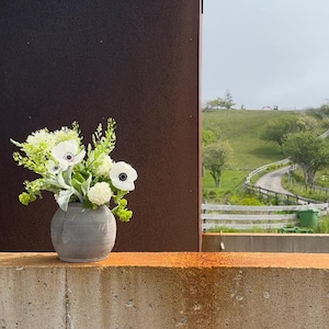 Puede incluir: Un ramo de anémonas blancas y vegetación en un jarrón de cerámica gris se encuentra sobre un borde de concreto con una mancha de óxido. El fondo muestra una colina verde cubierta de hierba con un camino de tierra sinuoso y una valla blanca.