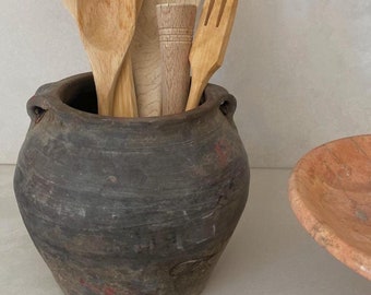 Könnte beinhalten: Eine Sammlung von Küchenutensilien aus Holz, darunter ein Löffel, ein Spatel und eine Gabel, werden in einem dunkelbraunen Keramiktopf mit strukturierter Oberfläche aufbewahrt.