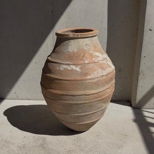 Op de afbeelding: Een grote, verweerde terracotta pot met een rond lichaam en een brede opening. De buitenkant is voorzien van horizontale ribbels rondom de pot, met vlekken van witte minerale afzettingen en een ruwe, aardse textuur op het oppervlak.