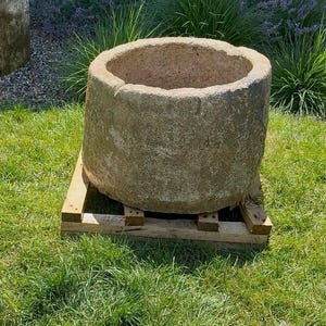 Puede incluir: Dos jardineras cilíndricas de piedra desgastada sobre césped. Una jardinera tiene una fuente. Las jardineras son de color marrón claro y tienen una textura rugosa. Las jardineras están sobre un palet de madera.