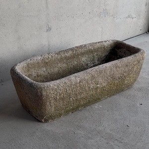 Puede incluir: Un abrevadero rectangular de piedra desgastada con una superficie rugosa y texturizada. El abrevadero es de un color gris verdoso apagado y parece estar hecho de piedra natural. La parte superior abierta sugiere que podría usarse para plantar o como elemento decorativo.
