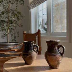 Peut inclure: Deux petits pots en terre cuite rustiques avec des anses posés sur une table ronde en bois. Les pots ont un glacis brun foncé et sont légèrement patinés.