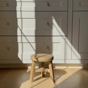 May include: A small, round wooden stool with three legs. The stool is made of light-colored wood and sits on a light-colored wooden floor. The background features white cabinets with gold-colored knobs. Sunlight casts shadows across the scene.