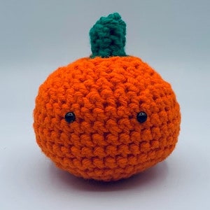 May include: A crocheted orange pumpkin with a green stem and two black bead eyes. The handmade pumpkin is a round shape and is likely a decorative item for the fall season or Halloween.