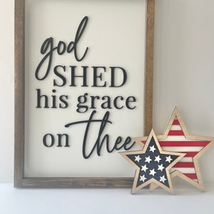 May include: A framed wooden sign with the words "god SHED his grace on thee" in black script. Two wooden star decorations, one with a blue star field and the other with red and white stripes, are next to the sign.