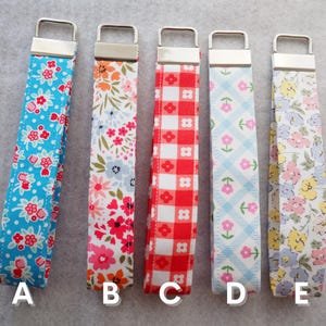 May include: Five fabric key fobs with silver-tone hardware. The patterns include a blue floral print, a floral print with pink and orange flowers, a red and white gingham check, a light blue floral pattern, and a yellow floral pattern. Each fob is labeled with a letter.