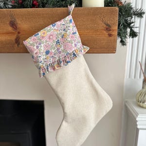 May include: A festive Christmas stocking with a cream-coloured body and a floral patterned cuff. The cuff displays pink, blue, and yellow flowers on a white background with a ruffled edge. The stocking hangs from a wooden mantel.