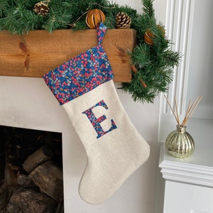 May include: A festive Christmas stocking in cream fabric, featuring a blue floral cuff and the letter 'E' appliquéd in the same pattern. It hangs from a wooden mantelpiece adorned with seasonal greenery and decorations.