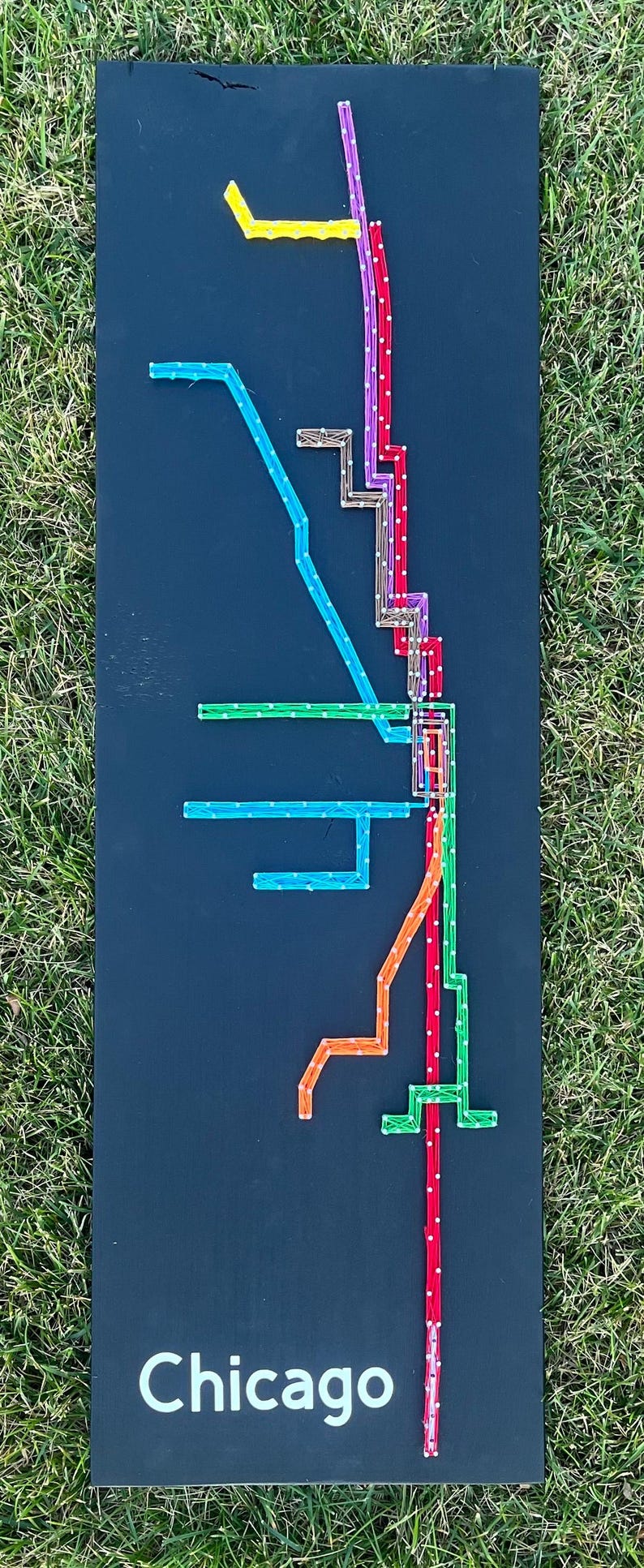 May include: A black background with a string art design of the Chicago 'L' train system. The lines are made of different colored strings, including red, orange, blue, green, purple, and yellow. The word "Chicago" is written in white at the bottom of the design.