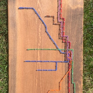 May include: A wooden sign with a string art depiction of the Chicago 'L' train system. The sign is stained brown and has the word "Chicago" written in white cursive lettering at the bottom.