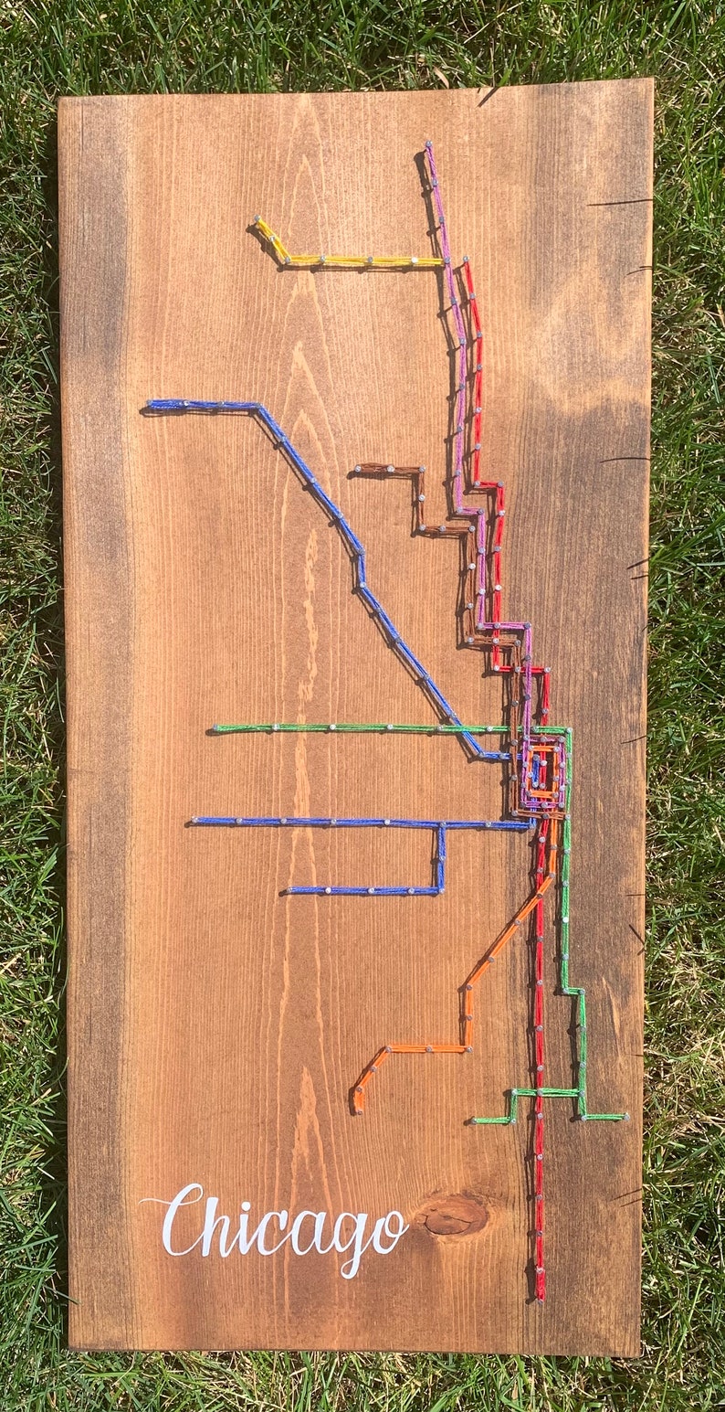 May include: A wooden sign with a string art depiction of the Chicago 'L' train system. The sign is stained brown and has the word "Chicago" written in white cursive lettering at the bottom.