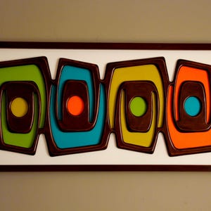 May include: Abstract wall art featuring four nested, geometric shapes in green, blue, yellow, and orange, each with a central circle. The shapes are outlined in dark brown and set against a white background, framed in a dark wood.