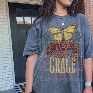Pode incluir: Uma camiseta cinza com uma estampa gr&aacute;fica de uma borboleta, flores e o texto "Amazing Grace 99 How Sweet The Sound".