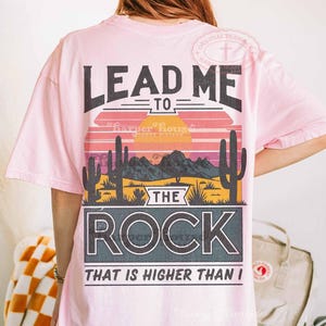 May include: Pink t-shirt with a graphic design that says "Lead Me To The Rock That Is Higher Than I". The graphic features a sunset scene with cacti and mountains.