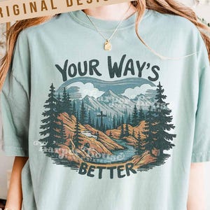 May include: A light green t-shirt with a graphic of a mountain landscape, river, and trees. The text "YOUR WAY'S BETTER" is above the landscape. The words "ORIGINAL DESIGNER!" are printed on a banner at the top.