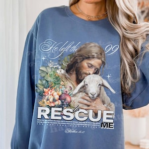 May include: A blue sweatshirt with a graphic print of Jesus holding a lamb. The graphic includes the text "He left the 99 to rescue me" and a Bible verse reference.