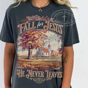 May include: Dark gray t-shirt with the text "Fall for Jesus" above a fall landscape scene with a church and tree. The text "He Never Leaves" is below the image. The shirt has a vintage design.