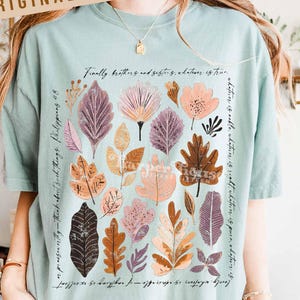 May include: A light teal t-shirt featuring a floral design with leaves in shades of purple, orange, and pink. The shirt has text that reads "Finally brothers and sisters, whatever is true." and "Harper House Three Sixteen."