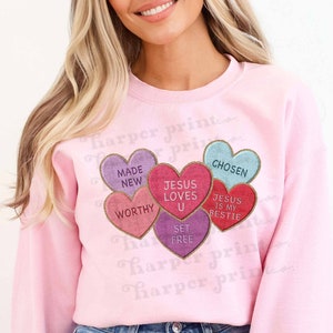 May include: Pink sweatshirt with a graphic of five candy conversation hearts. The hearts say "Made New", "Worthy", "Jesus Loves U", "Set Free", and "Jesus is My Bestie".