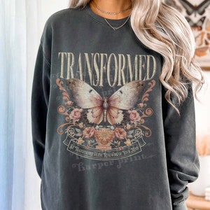 Op de afbeelding: Een donkergrijs sweatshirt met een vintage-geïnspireerd ontwerp met een vlinder met bloemmotieven. De tekst "TRANSFORMED" is in vetgedrukte letters boven de vlinder gedrukt. De tekst "BE TRANSFORMED BY THE RENEWING OF YOUR MIND" is onder de vlinder gedrukt.