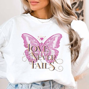 Può includere: Felpa bianca a maniche lunghe con una grafica di farfalla rosa e il testo "LOVE NEVER FAILS" in lettere dorate. La farfalla ha un cuore al centro con il testo "CORINTHIANS 13:8".
