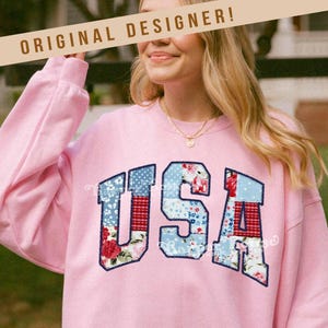 May include: A pink sweatshirt with the word "USA" in large, patchwork letters. The letters are made up of different fabrics with floral, gingham, and blue patterns. The sweatshirt has a crew neck and long sleeves.