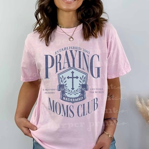 Può includere: Maglietta rosa con un aspetto vissuto che presenta una grafica blu e bianca con il testo "Established 2003 Praying Redeemed Moms Club". La grafica include una croce e una ghirlanda.