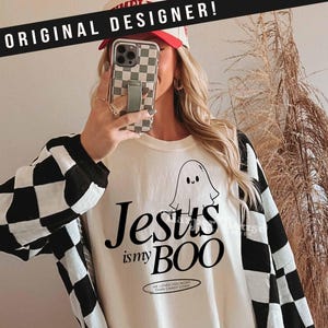 May include: Cream-colored long-sleeve shirt with black and white checkered sleeves. The shirt features the text "Jesus is my BOO" with a ghost graphic. The phrase "He loves you more than candy corn" is also printed on the shirt.