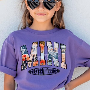 May include: Purple short-sleeved t-shirt with a floral patchwork design that spells out "MINI PRAYER WARRIOR".