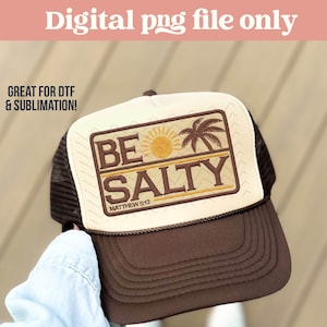 May include: A brown and cream-colored trucker hat with the words "BE SALTY" and "MATTHEW 5:13" printed on the front. The design includes a sun and palm tree graphic. The hat has a mesh back and a curved brim.