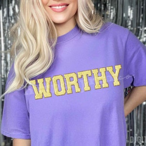 May include: A purple t-shirt with the word "WORTHY" in gold glitter letters.