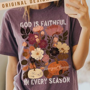 May include: Purple t-shirt with a floral design and the text "GOD IS FAITHFUL IN EVERY SEASON." The design includes flowers and pumpkins in shades of pink, orange, and white. The shirt also has the text "ORIGINAL DESIGNER!" and "Harper House THREE SIXTEEN."