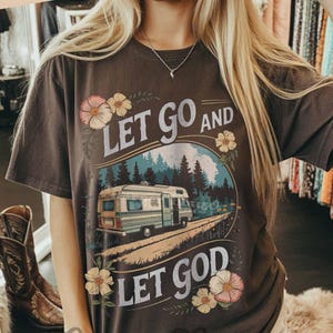 May include: A dark brown t-shirt with the text "LET GO AND LET GOD" and a graphic of a camper van on a road in front of a mountain and forest scene. The shirt has floral accents and the words "ORIGINAL DESIGNER!" at the top.