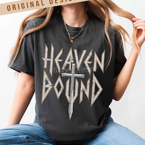 May include: Dark gray t-shirt with the words "HEAVEN BOUND" in a distressed, white, gothic-style font, with a silver cross graphic. The shirt also features the text "ORIGINAL DESIGNER!" at the top.