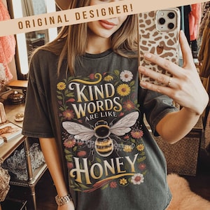Peut inclure: T-shirt gris fonc&eacute; avec un motif floral et une abeille. Le t-shirt pr&eacute;sente le texte "Kind Words Are Like Honey" dans une police d&eacute;corative. Le motif comprend une grande abeille avec des ailes d&eacute;taill&eacute;es et des fleurs color&eacute;es.