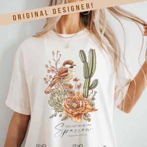 May include: A white t-shirt with a brown sparrow perched on a cactus and flowers. The text "His eye is on the Sparrow Matthew 10:29-31" is printed on the shirt. The text "harper house THREE SIXTEEN" is printed below the sparrow.
