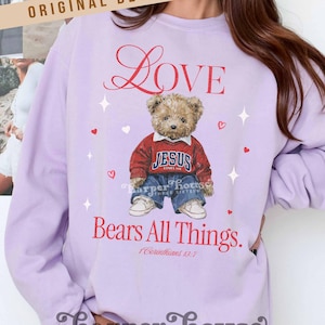 May include: A light purple sweatshirt with the word "Love" in red script. It features a teddy bear wearing a red sweater with "JESUS LOVES ME" and blue jeans. The text "Bears All Things" and "1 Corinthians 13:7" are also printed on the sweatshirt.