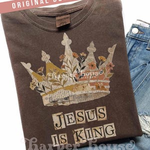 Puede incluir: Una camiseta marrón con un diseño de corona con detalles florales y el texto "JESUS IS KING". La corona es un collage de papel. La camiseta tiene un aspecto vintage. Las palabras "ORIGINAL DESIGNER!" están en la parte superior.