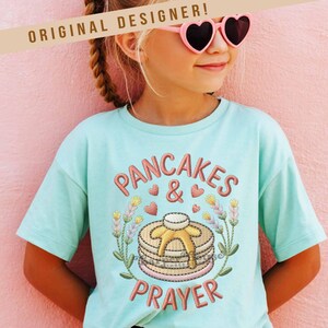 May include: A light blue t-shirt with the words "PANCAKES & PRAYER" embroidered in a reddish-brown color. The design includes a stack of pancakes with syrup, hearts, and floral accents. The text "ORIGINAL DESIGNER!" is above the shirt.