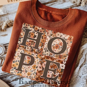 May include: A rust-colored sweatshirt with the word "HOPE" in large, dark brown letters. The letters are filled with a floral pattern in shades of orange, brown, and cream. The sweatshirt is folded neatly, and the text "ORIGINAL DESIGNER!" is visible.