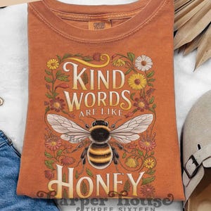 Peut inclure: Un t-shirt orange avec le texte "Kind Words Are Like Honey" et une illustration d&eacute;taill&eacute;e d'une abeille. Le motif est entour&eacute; d'&eacute;l&eacute;ments floraux. Le t-shirt a un col rond et des manches courtes.