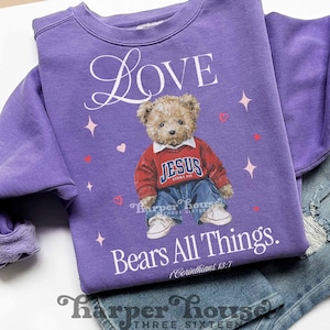 Puede incluir: Una sudadera morada con la palabra "Love" en escritura blanca. Presenta un oso de peluche con un suéter rojo con "JESUS LOVES ME" y jeans azules. El texto "Bears All Things" y "1 Corinthians 13:7" también están impresos.